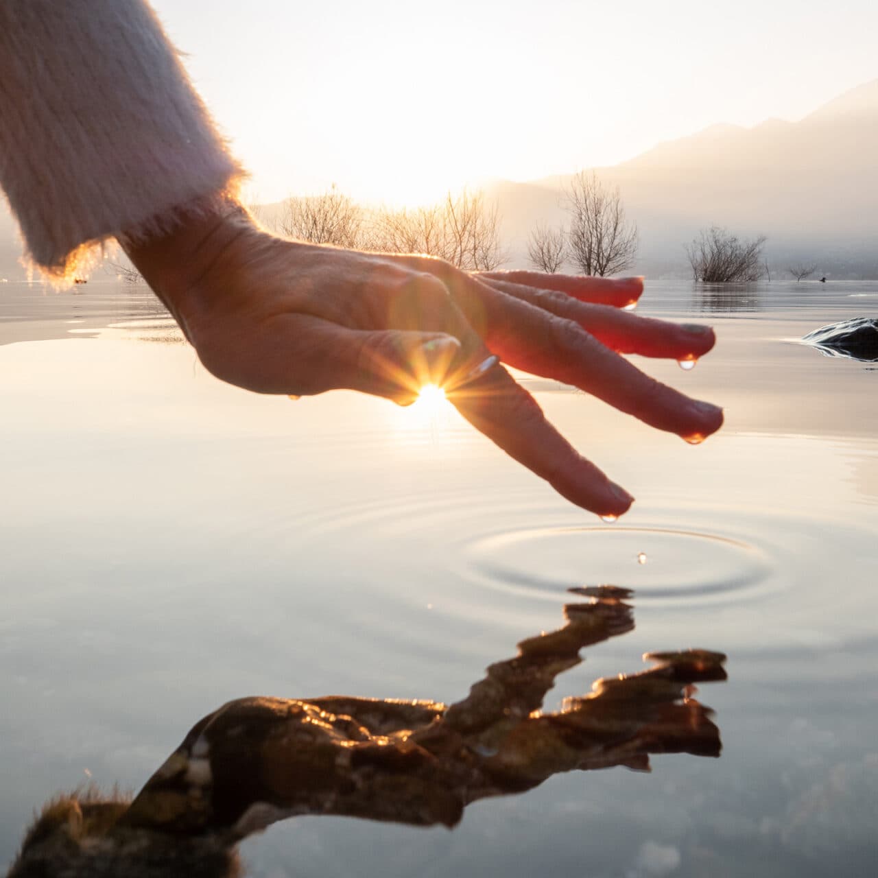 Detail of hand touching and caressing water surface of beautiful lake at sunset, mountain view. ...