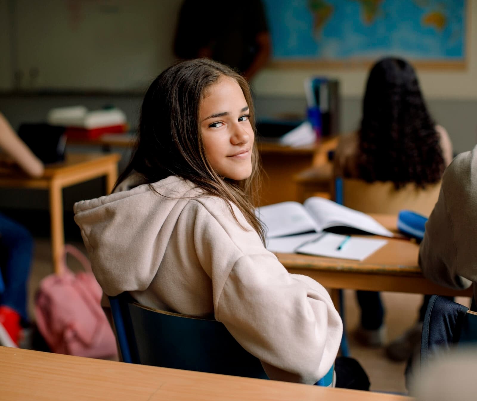 Bilden visar en klassrumsmiljö där en ung flicka sitter vid en skolbänk och tittar tillbaka mot kameran med ett halvt leende. Hon bär en beige huvtröja och hennes hår är löst. I bakgrunden kan man se andra elever som är fokuserade på sina skolarbeten och en världskarta som är upphängd på väggen, vilket antyder att det kan vara en geografilektion. Det finns böcker och skrivmaterial på borden, och atmosfären verkar lugn och studieinriktad.