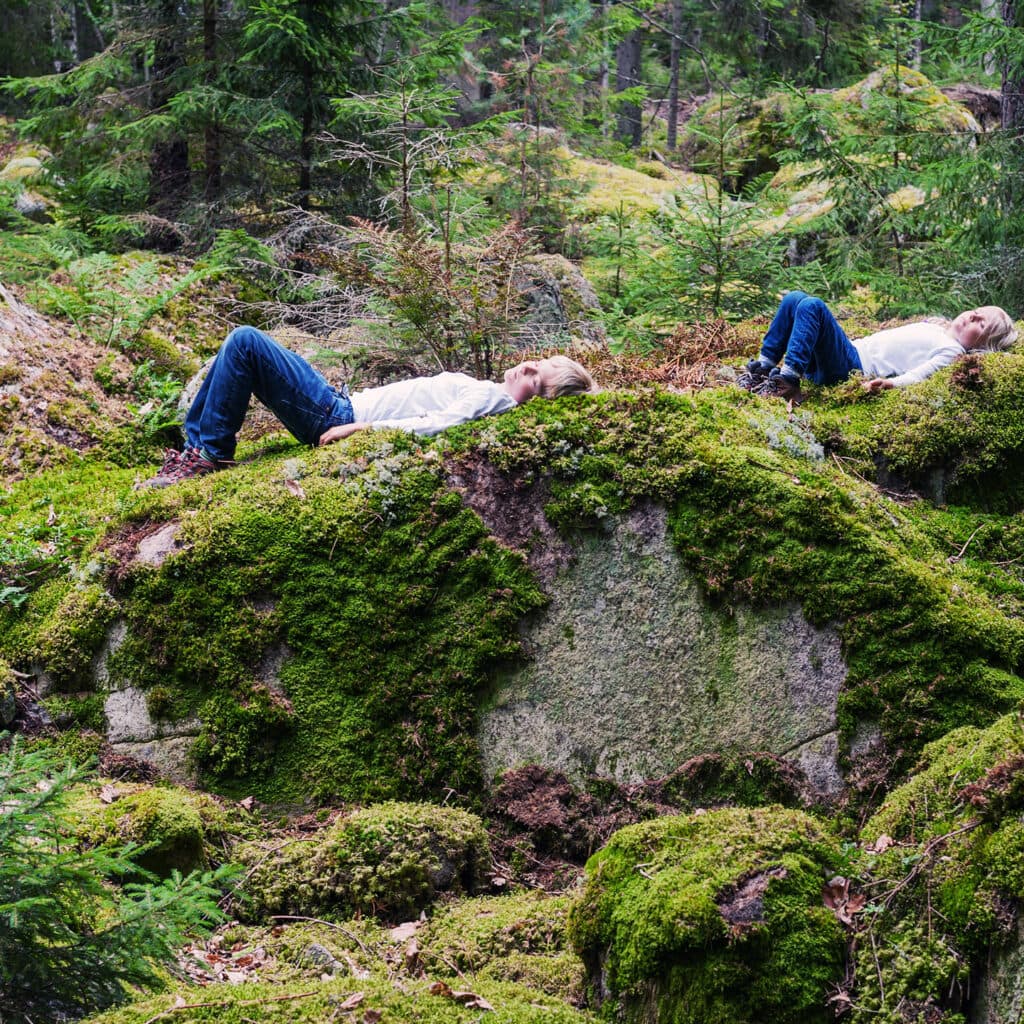 Två barn vilar på stora mossiga stenar i en skog.