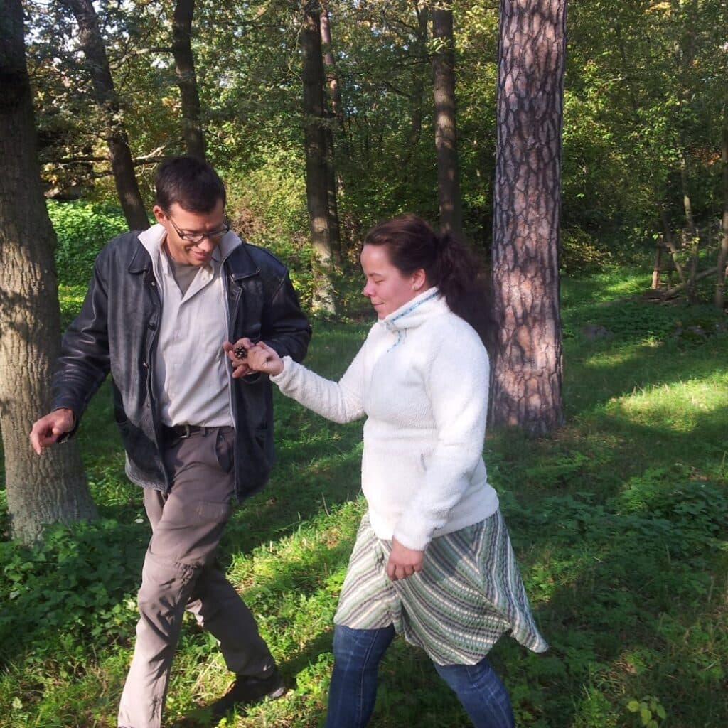 Bilden visar två personer som går tillsammans i en skog eller park. De verkar vara engagerade i en konversation eller aktivitet, och omgivningen är grön och lummig med träd och gräs.