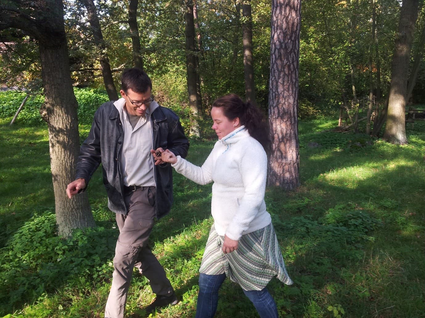 Bilden visar två personer som går tillsammans i en skog eller park. De verkar vara engagerade i en konversation eller aktivitet, och omgivningen är grön och lummig med träd och gräs.