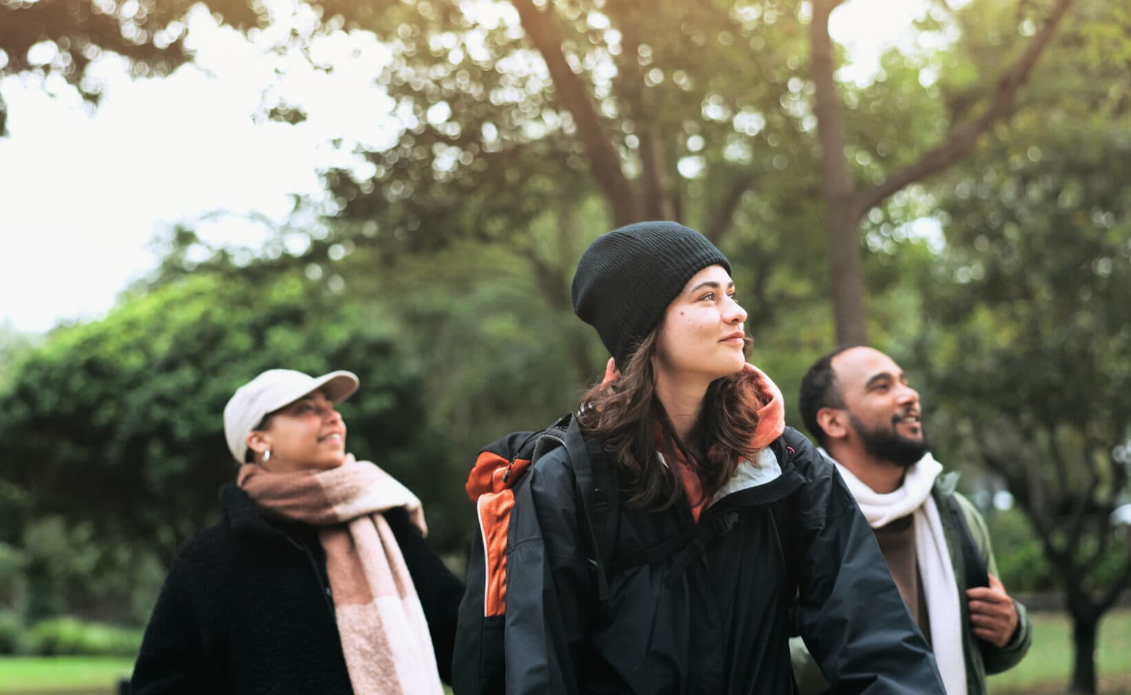 Bilden visar tre personer som promenerar utomhus i en park. De ser glada ut och är klädda i varma kläder. I bakgrunden syns träd och grönska. Det verkar vara en solig dag.
