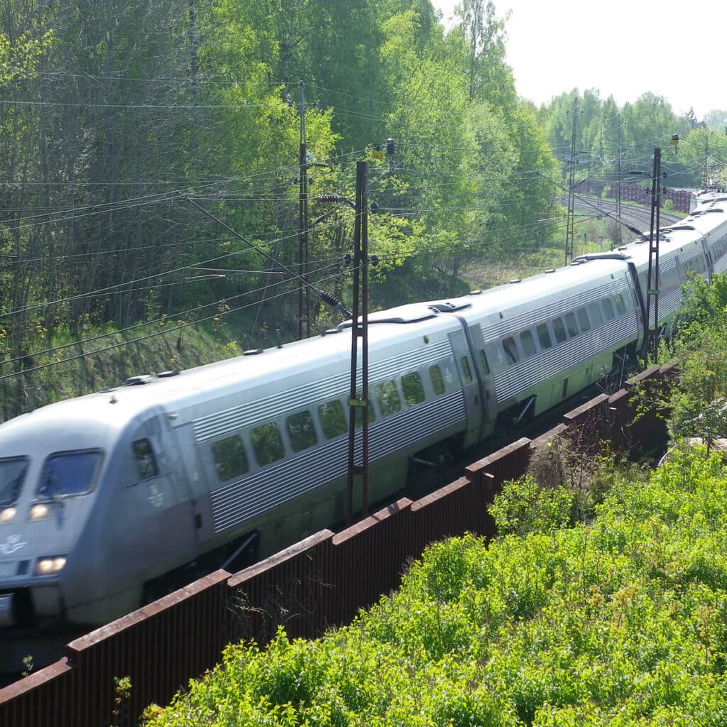Bilden visar ett tåg som kör på en järnväg genom ett grönskande landskap. Tåget är långt och omges ...