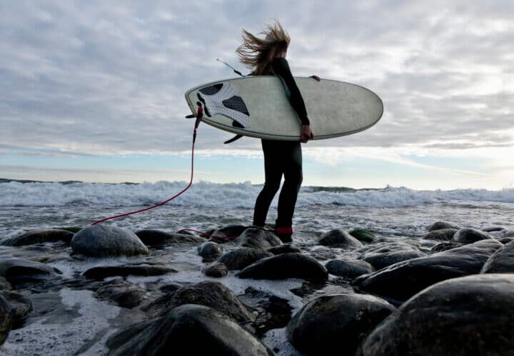 Person med surfbräda vid en stenig strand som visar hur strandskydd i Sverige gör stränder ...