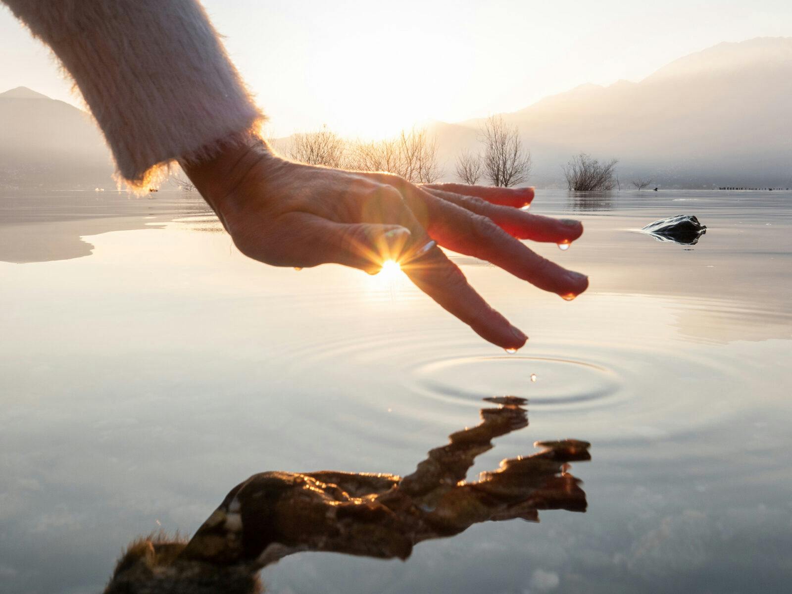 Detail of hand touching and caressing water surface of beautiful lake at sunset, mountain view. Purity freshness clean concept, one person touching lake with hand